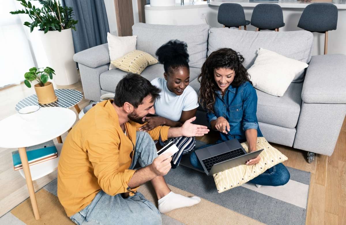 Three excited friends buying a vacation tickets trip online with a credit card and a laptop sitting on a floor in the living room at home