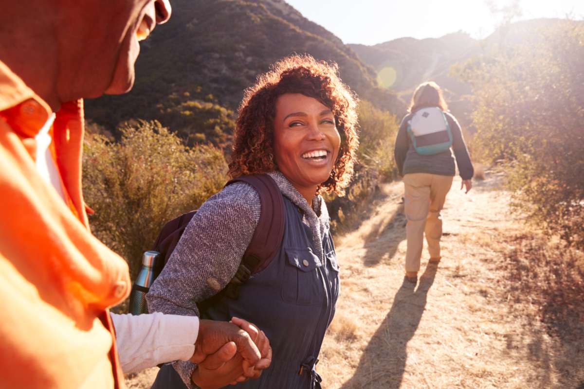 Woman Helping Man On Trail As Group Of Senior Friends Go Hiking In Countryside Together